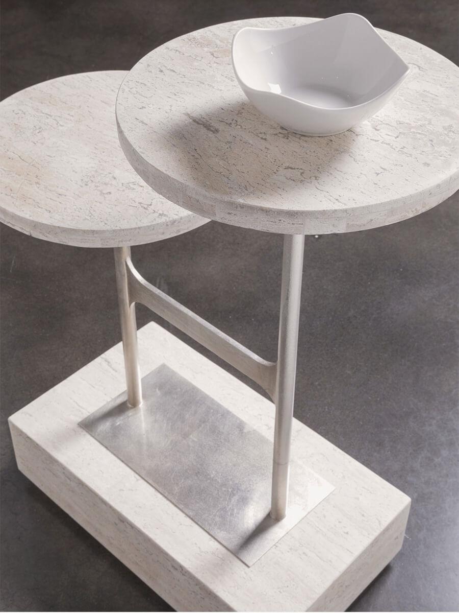 A travertine looking accent table with two tiers connected with a metal brace on a large slab of travertine looking material.